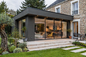 Extension moderne en verre d'une maison en Île-de-France avec portes coulissantes, rattachée à une maison en pierre, s'ouvrant sur un patio carrelé et un jardin paysager. L'espace intérieur comprend des sièges et des salles à manger visibles.
