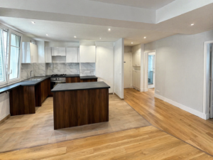 Cuisine moderne avec des armoires en bois foncé, des comptoirs noirs, des armoires supérieures blanches et un dosseret en marbre. L'agencement ouvert est relié à un espace de vie avec un sol en bois clair et de grandes fenêtres apportant de la lumière naturelle.