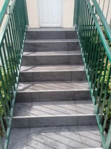 Escalier en pierre grise avec rampe verte menant à une porte blanche.