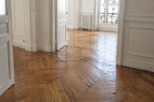 Un appartement vide et ensoleillé, avec des murs et des portes blancs, des sols en bois à chevrons provenant d'une pose de parquet et d'une rénovation de parquet ancienne, de grandes fenêtres et un radiateur sous une fenêtre.