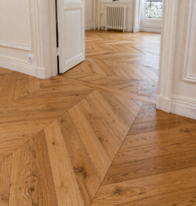 Les pièces adjacentes sont traversées par des planchers en bois à motifs chevron, de couleur marron clair, issus d'une récente rénovation des sols (carrelage, parquet, stratifié). Des portes blanches, des murs avec des moulures, un radiateur et une fenêtre avec un garde-corps en fer forgé complètent l'ensemble.