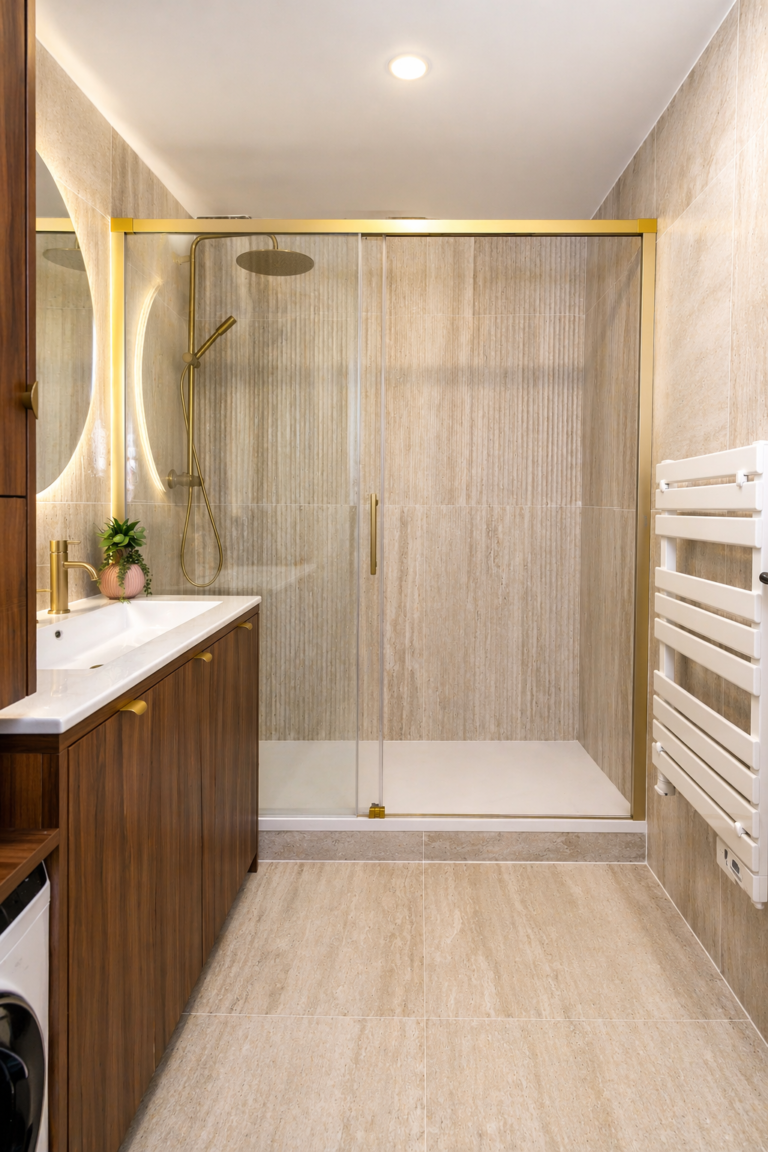 Salle de bains moderne avec carrelage beige, cabine de douche en verre avec accessoires dorés, meuble-lavabo en bois, miroir rond rétro-éclairé, petite plante en pot et sèche-serviettes blanc fixé au mur.