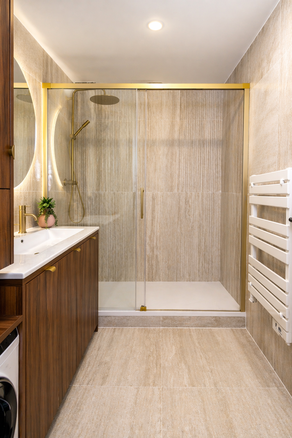 Salle de bains moderne avec carrelage beige, cabine de douche en verre avec accessoires dorés, meuble-lavabo en bois, miroir rond rétro-éclairé, petite plante en pot et sèche-serviettes blanc fixé au mur.