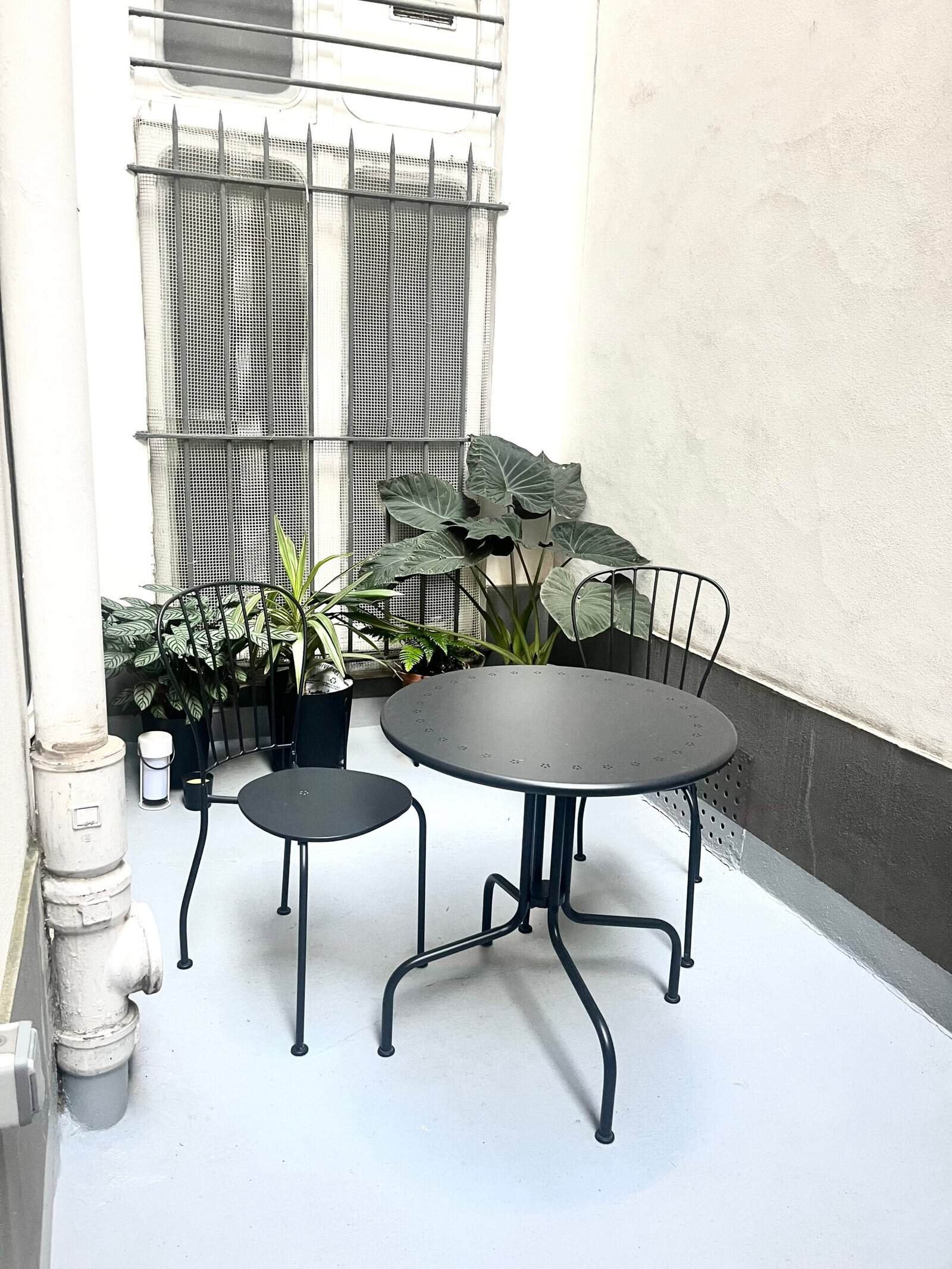 Balcon avec table ronde et chaises en métal, décor végétal et ambiance urbaine.